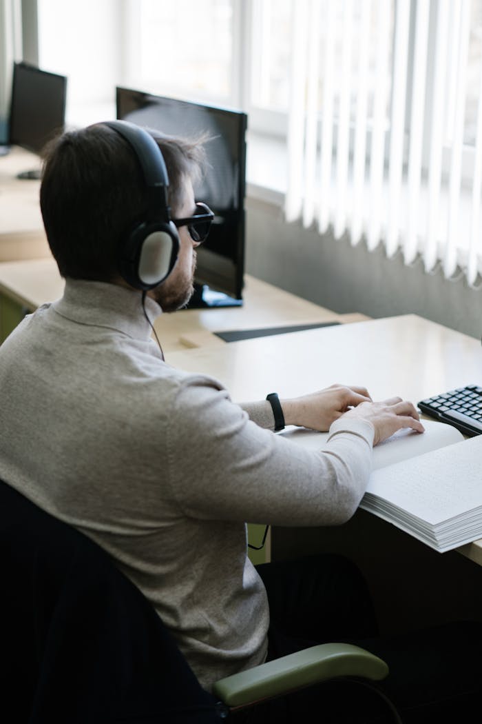 A visually impaired man reading a braille book indoors with headphones on.